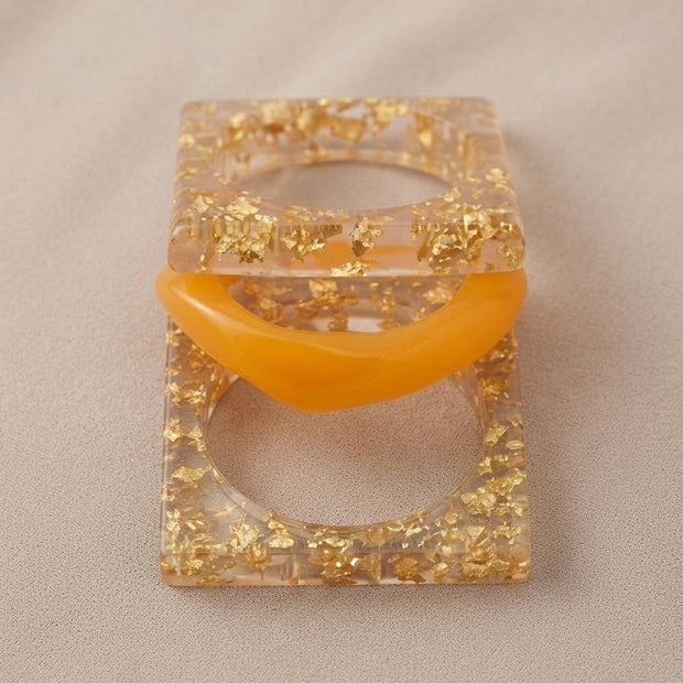 Top view of square gold foil resin bangles and amber dome bangle stacked flat on beige surface
