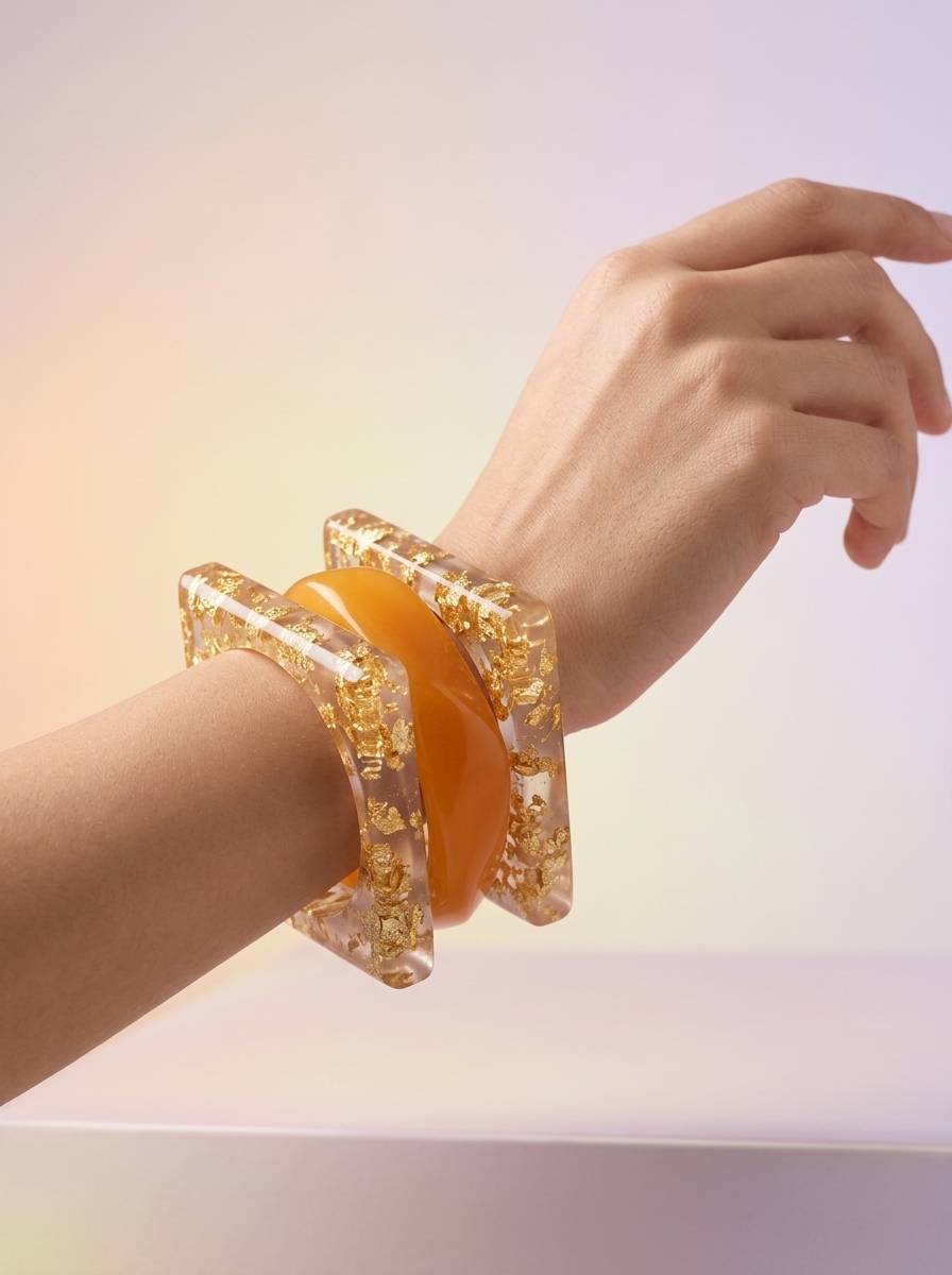 Square gold foil resin bangles and amber bangle worn stacked on a woman's wrist against pastel background