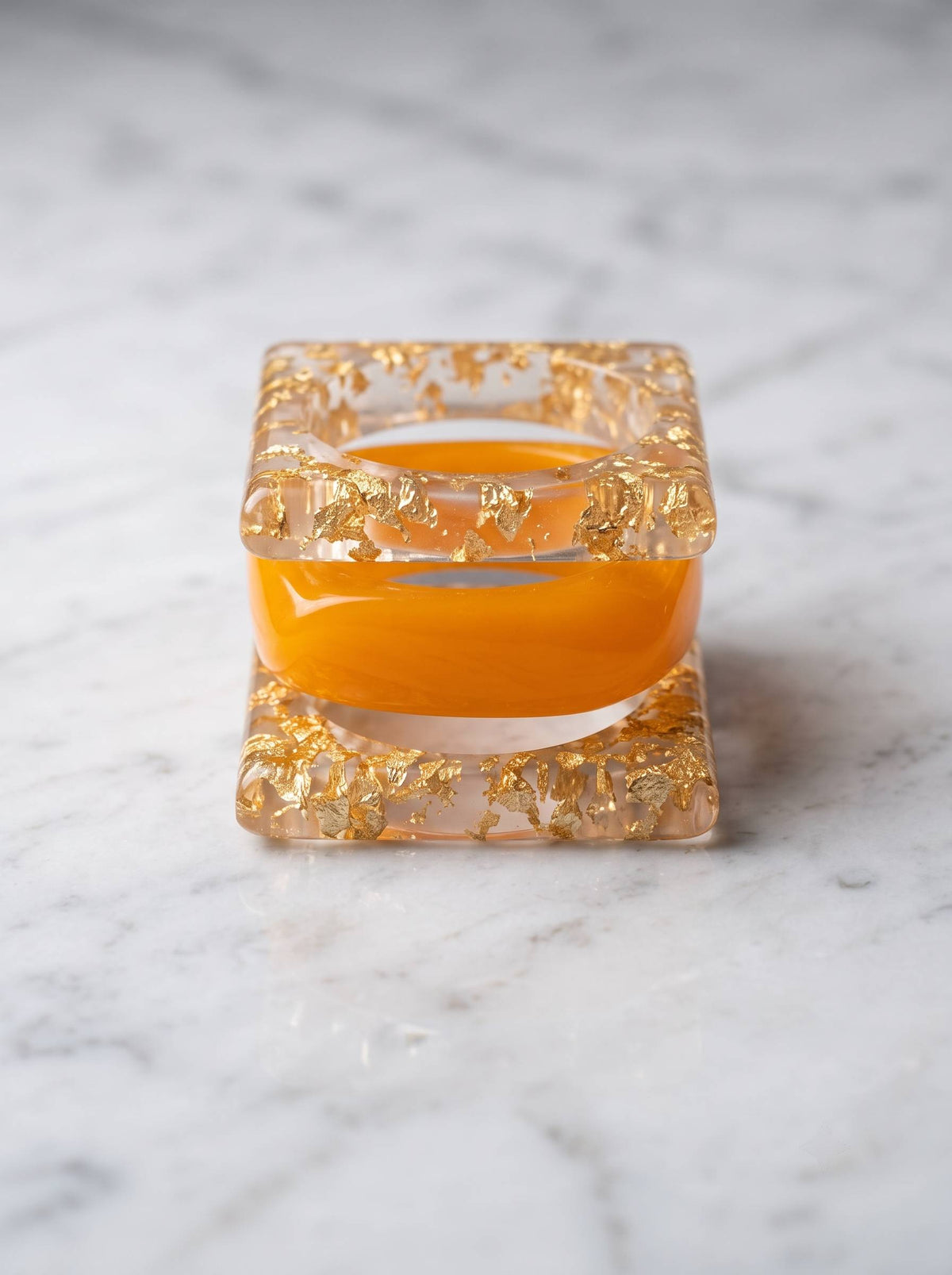 Front-facing view of square gold foil resin bangle and amber dome bangle nested together on marble surface