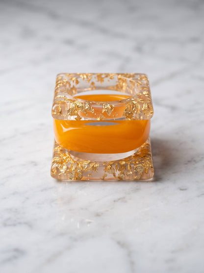 Front-facing view of square gold foil resin bangle and amber dome bangle nested together on marble surface