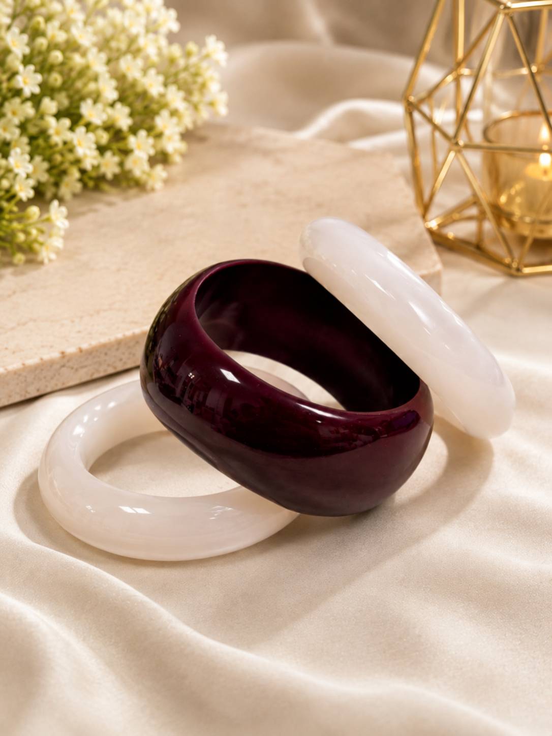 Two white resin bangles and one dark burgundy resin kada laid flat on cream satin fabric