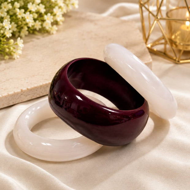 Two white resin bangles and one dark burgundy resin kada laid flat on cream satin fabric