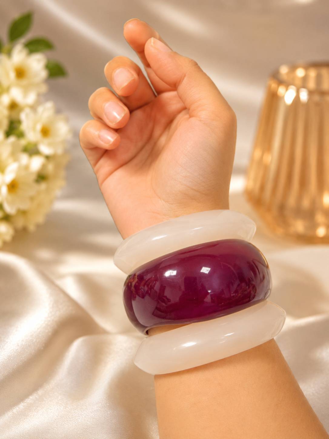Woman wearing stacked white and burgundy resin bangles on her wrist against a soft draped background