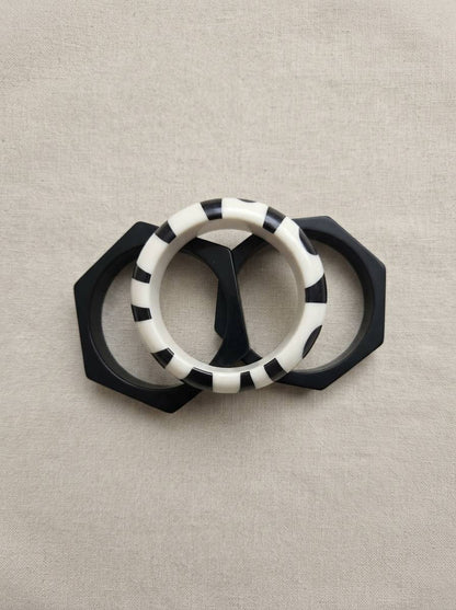 Flat lay of two black octagonal bangles and one round black and white printed resin bangle on grey linen