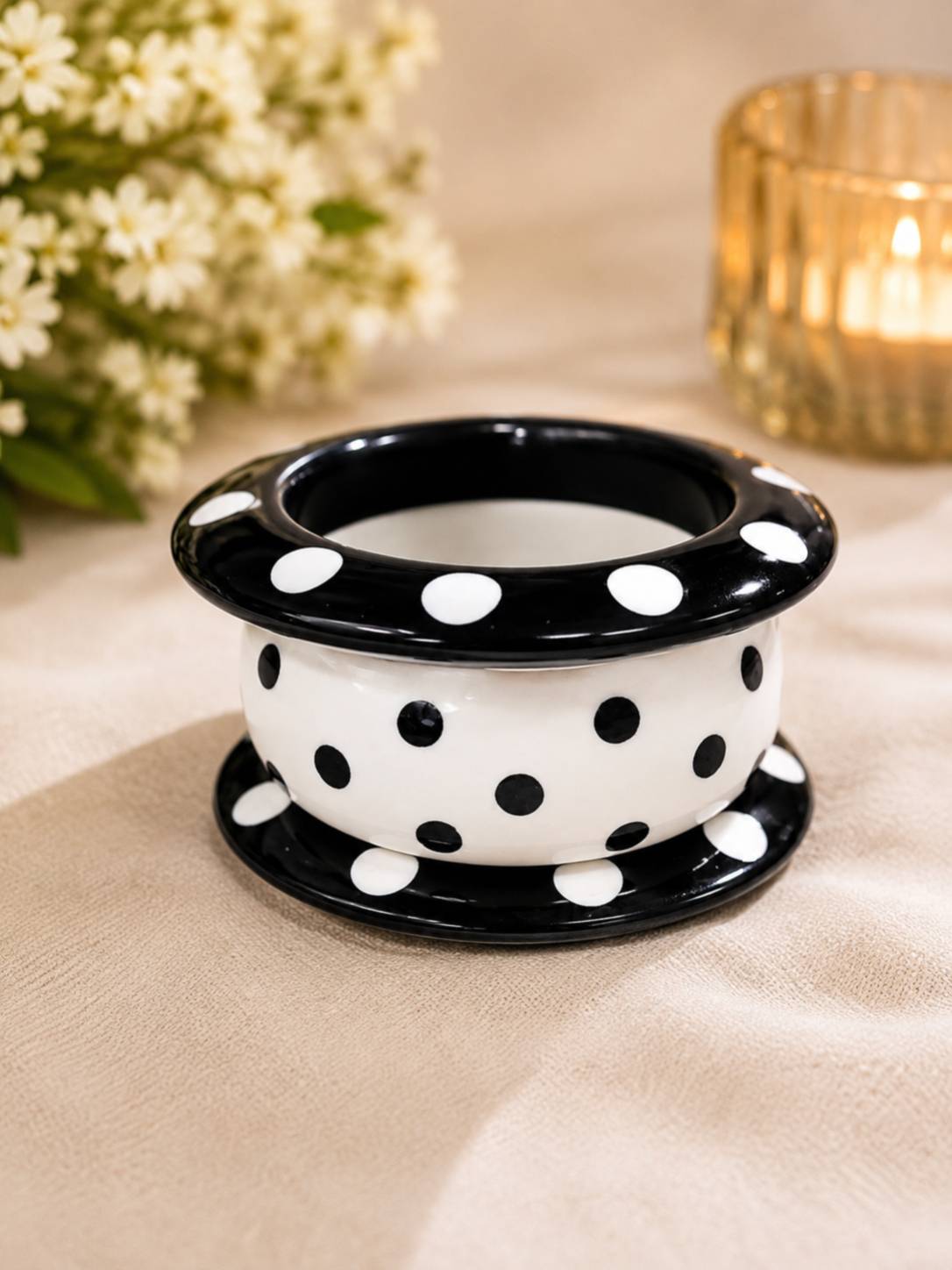 Three stacked black and white polka dot resin bangles viewed from above on a beige fabric surface with flowers and a candle in the background