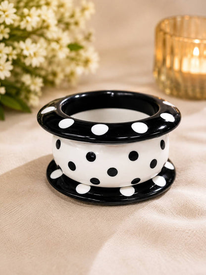 Three stacked black and white polka dot resin bangles viewed from above on a beige fabric surface with flowers and a candle in the background
