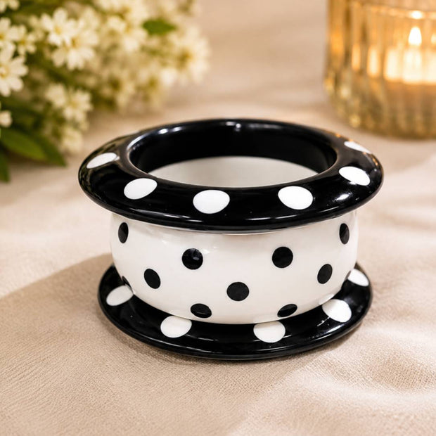 Three stacked black and white polka dot resin bangles viewed from above on a beige fabric surface with flowers and a candle in the background