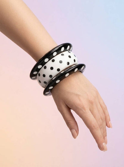 Woman wearing all three black and white polka dot resin bangles stacked on her wrist against a pastel pink and purple gradient background
