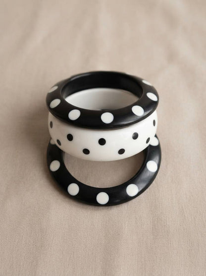 Three polka dot resin bangles stacked flat on a beige linen surface showing two black bangles with white dots and one white bangle with black dots