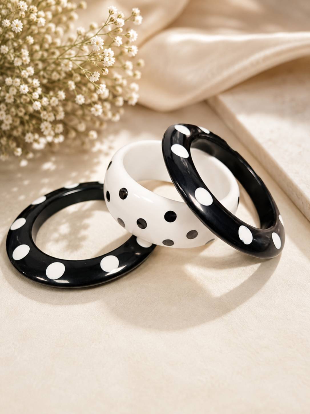 Three black and white polka dot resin bangles laid flat and overlapping on a cream fabric surface next to baby's breath flowers and a silk drape