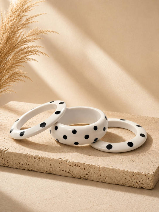 Three white polka dot resin bangles arranged on a sandy stone surface with dried pampas grass in the background