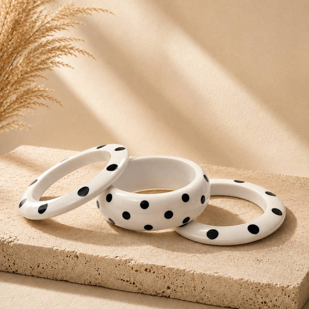 Three white polka dot resin bangles arranged on a sandy stone surface with dried pampas grass in the background