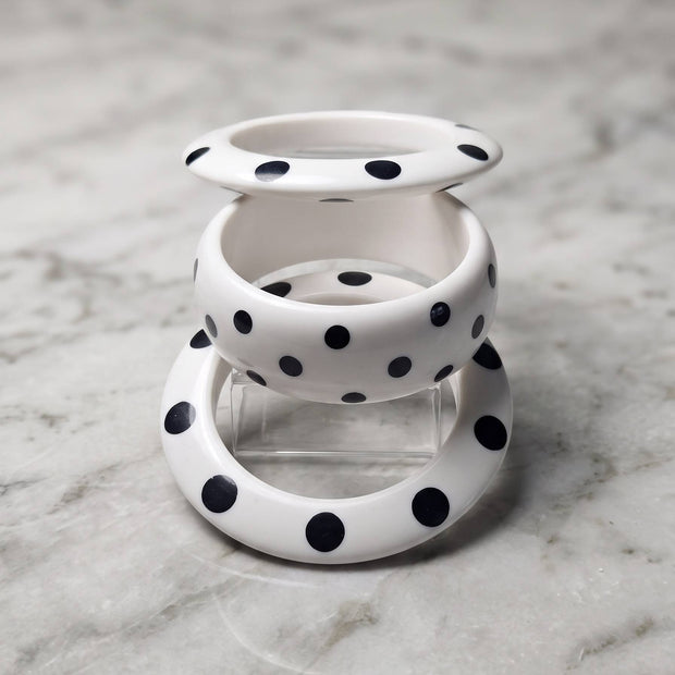 Three white resin bangles with black polka dots stacked vertically on a clear stand against a marble surface