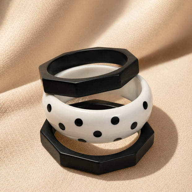 Three resin bangles stacked on beige fabric — two black octagonal and one white polka-dot bangle