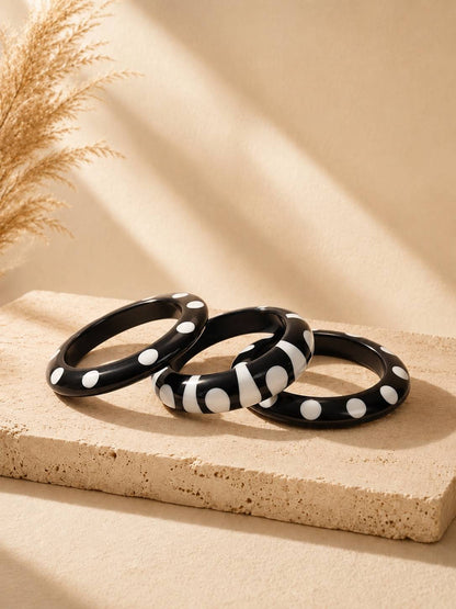 Three black and white resin bangles with polka dot and zebra stripe patterns arranged on a beige stone surface with dried pampas grass