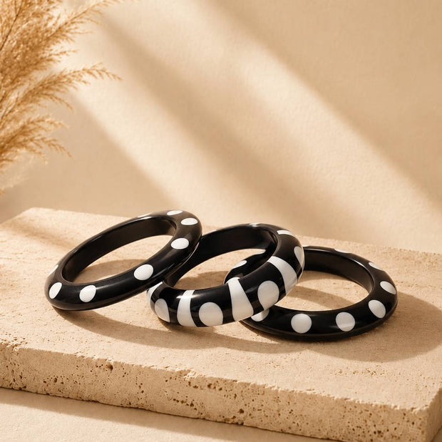 Three black and white resin bangles with polka dot and zebra stripe patterns arranged on a beige stone surface with dried pampas grass