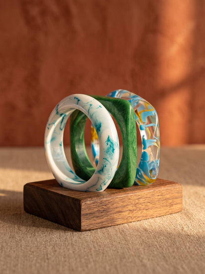 White teal, solid green, and blue yellow resin bangles displayed upright on a small wooden block