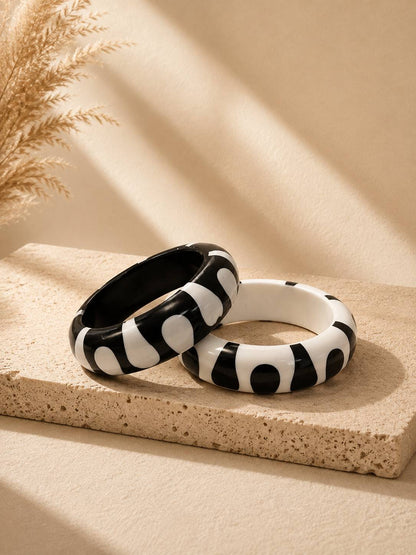 Two black and white resin bangle kadas with abstract splodge pattern resting on a sandy stone tray with dried pampas grass in the background