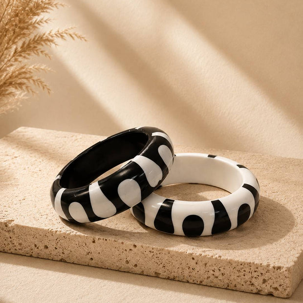 Two black and white resin bangle kadas with abstract splodge pattern resting on a sandy stone tray with dried pampas grass in the background