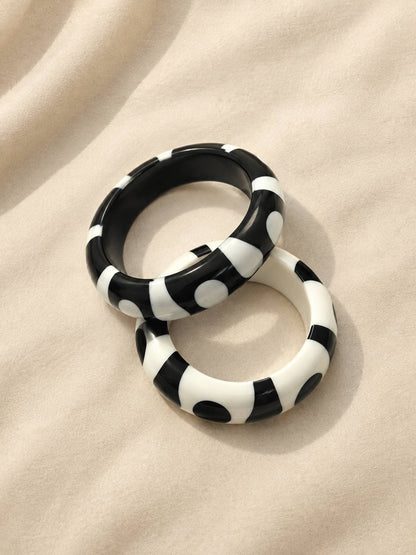 Black-base and white-base resin bangles with contrasting dot and stripe pattern placed side by side on a soft cream fabric surface