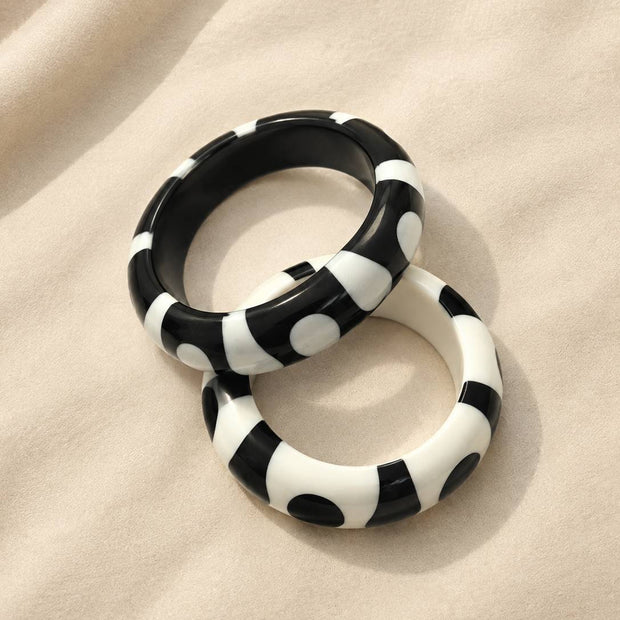 Black-base and white-base resin bangles with contrasting dot and stripe pattern placed side by side on a soft cream fabric surface