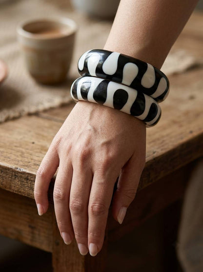 Both black and white resin bangle kadas stacked together on a woman's wrist resting on a wooden table with a ceramic cup in the background