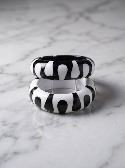 Two resin bangles with bold black and white graphic pattern stacked vertically on a white marble surface showing their rounded profile