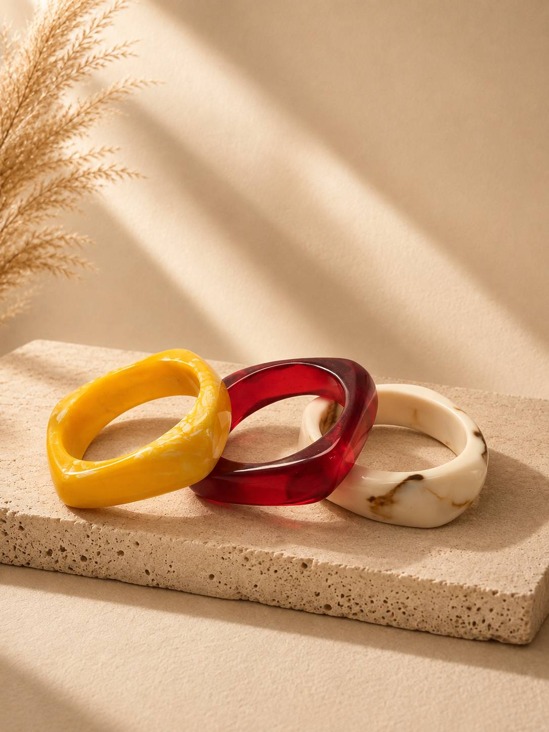 Three chunky resin bangles in yellow marble, translucent ruby red and ivory marble arranged on a textured stone surface