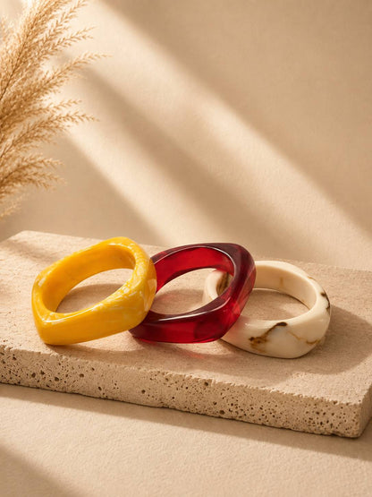 Three chunky resin bangles in yellow marble, translucent ruby red and ivory marble arranged on a textured stone surface