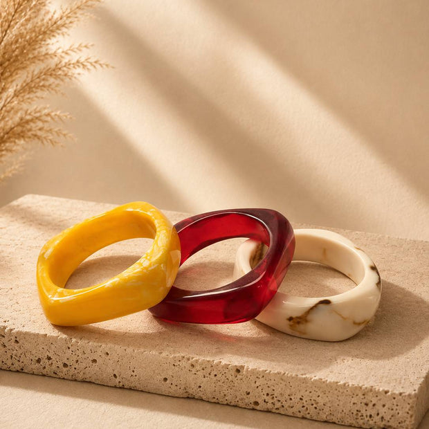 Three chunky resin bangles in yellow marble, translucent ruby red and ivory marble arranged on a textured stone surface
