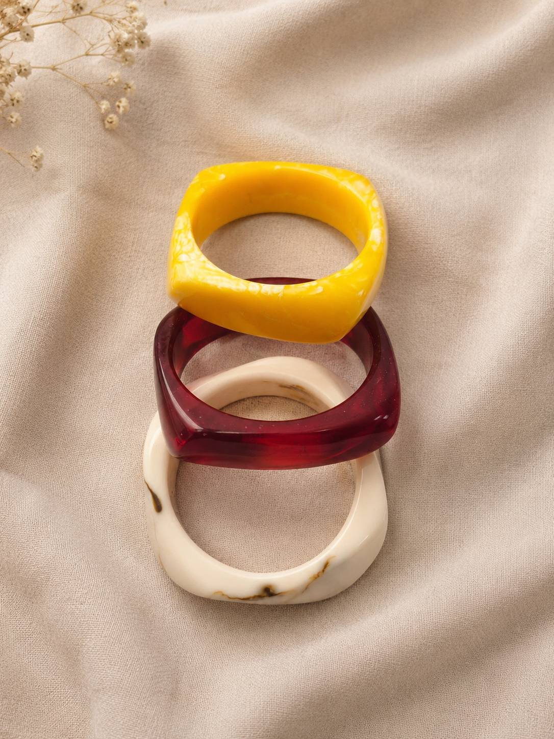Stack of three glossy resin bangles in mustard yellow, deep red and cream marble laid on draped beige fabric