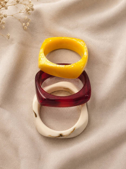 Stack of three glossy resin bangles in mustard yellow, deep red and cream marble laid on draped beige fabric