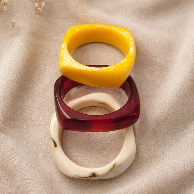 Stack of three glossy resin bangles in mustard yellow, deep red and cream marble laid on draped beige fabric