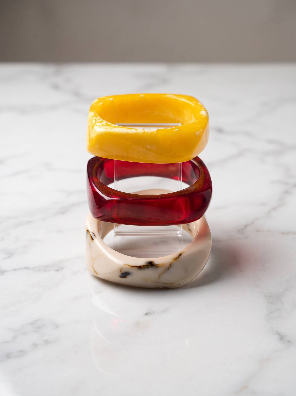 Three chunky resin bangles stacked vertically on a white marble surface showing yellow opaque, translucent red and ivory marble finishes