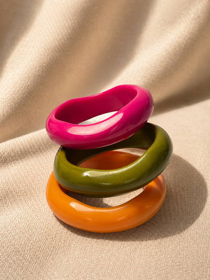 Three glossy resin bangles in pink, olive green and orange stacked upright on a beige fabric surface