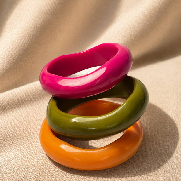 Three glossy resin bangles in pink, olive green and orange stacked upright on a beige fabric surface