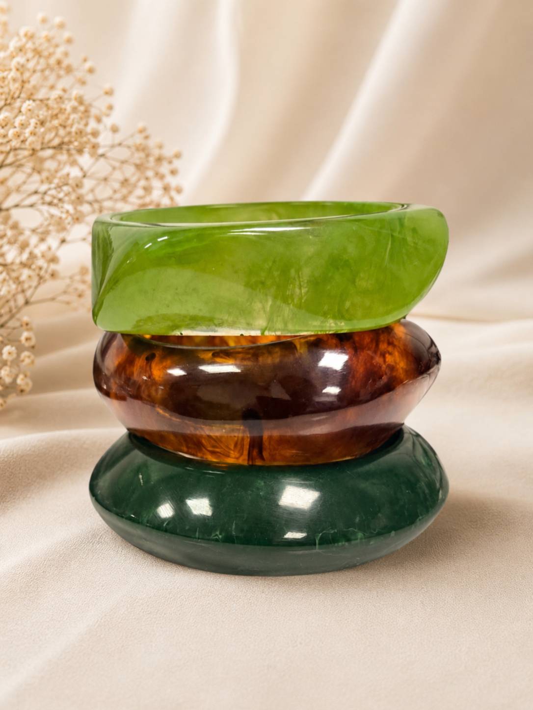 Three stacked resin bangles in lime green, tortoiseshell, and forest green displayed on white fabric with dried flowers
