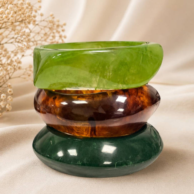 Three stacked resin bangles in lime green, tortoiseshell, and forest green displayed on white fabric with dried flowers
