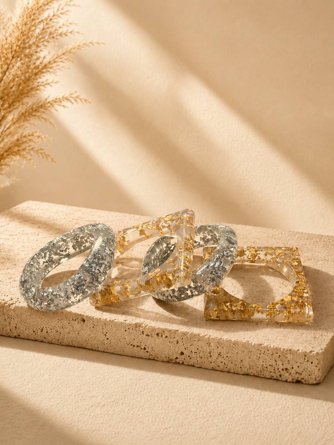 Four clear resin bangles with gold and silver foil, two round and two square, displayed on a stone surface with dried pampas grass