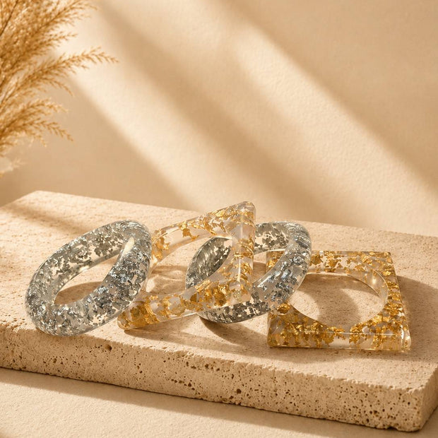 Four clear resin bangles with gold and silver foil, two round and two square, displayed on a stone surface with dried pampas grass