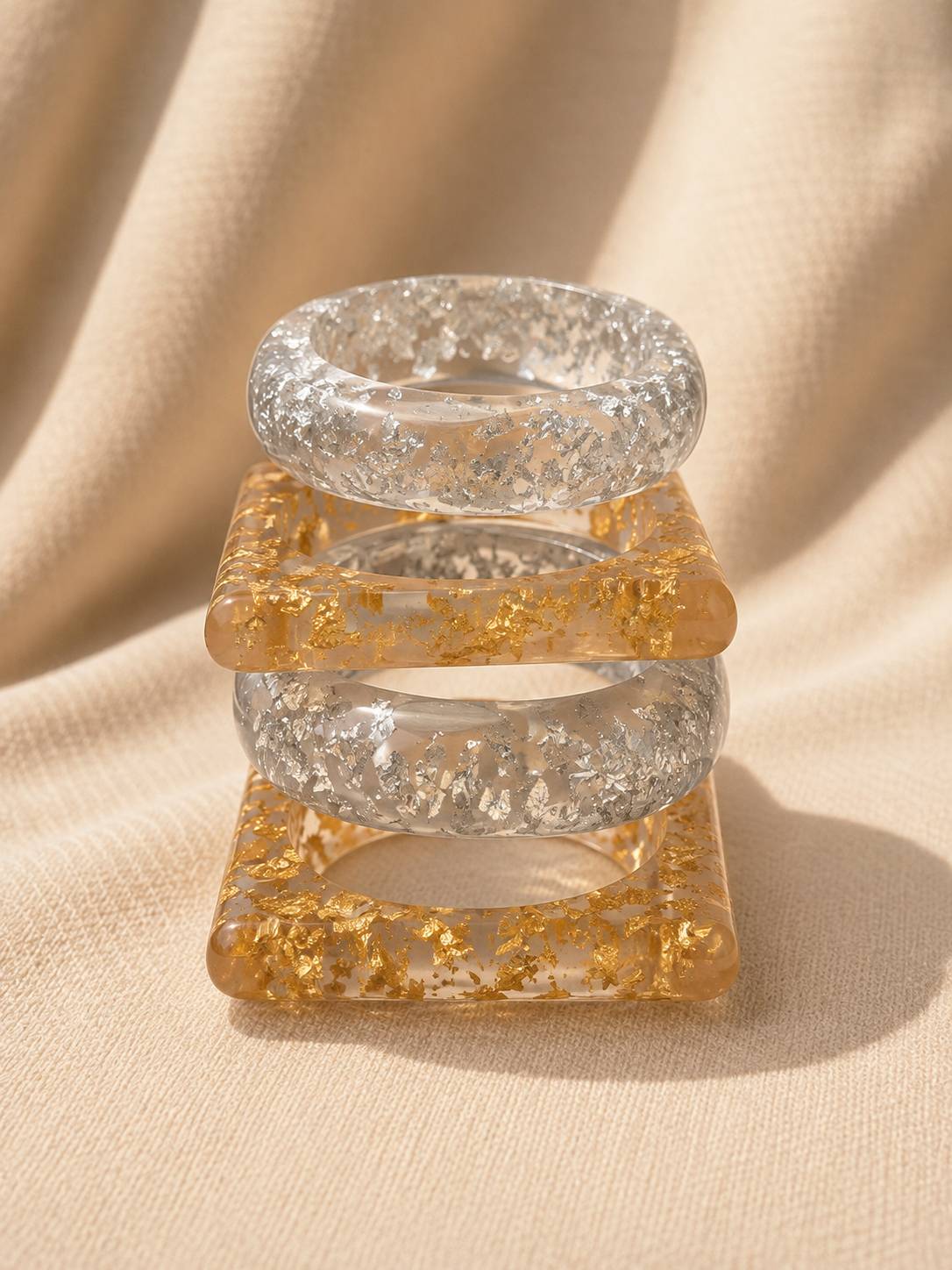 Stack of four clear resin bangles alternating round silver foil and square gold foil designs, stacked vertically on cream fabric