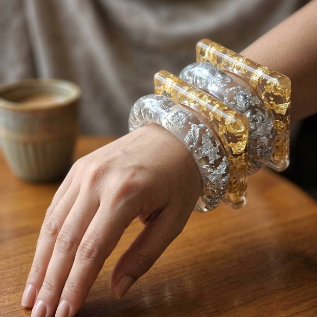 Woman wearing all four gold and silver foil resin bangles stacked on her wrist, resting hand on wooden table beside a coffee cup