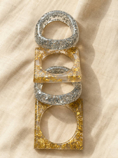 Four resin bangles laid flat in a vertical row on linen fabric, alternating between silver foil round bangles and gold foil square bangles