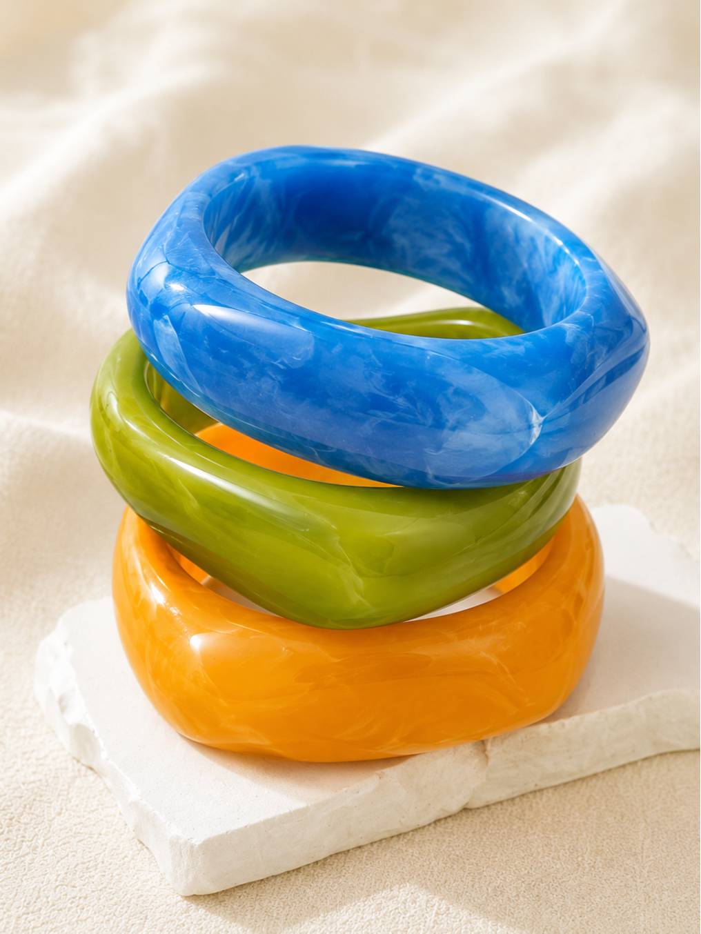 Three glossy resin marble bangles in blue, green, and orange stacked on a white stone block