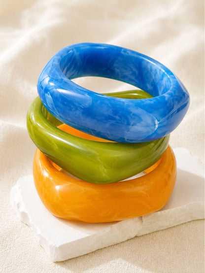Three glossy resin marble bangles in blue, green, and orange stacked on a white stone block