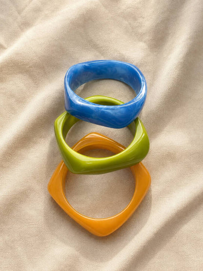 Blue, green, and orange angular geometric resin bangles stacked flat on a beige fabric surface