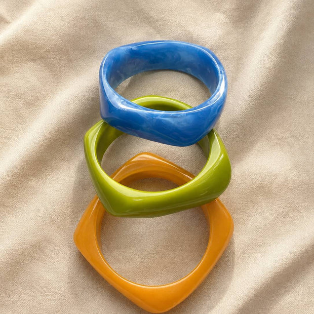Blue, green, and orange angular geometric resin bangles stacked flat on a beige fabric surface