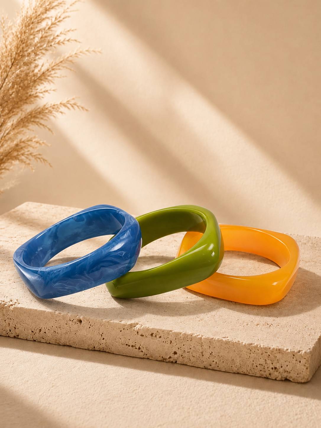 Three square-shaped resin bangles in blue, green, and orange displayed side by side on a stone slab