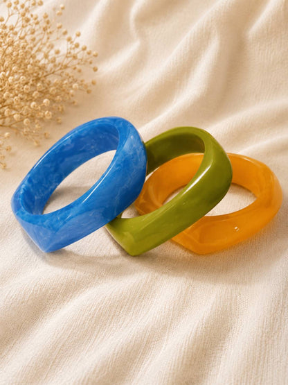 Three angular resin bangles in blue, green, and orange arranged in a fan layout on white cloth