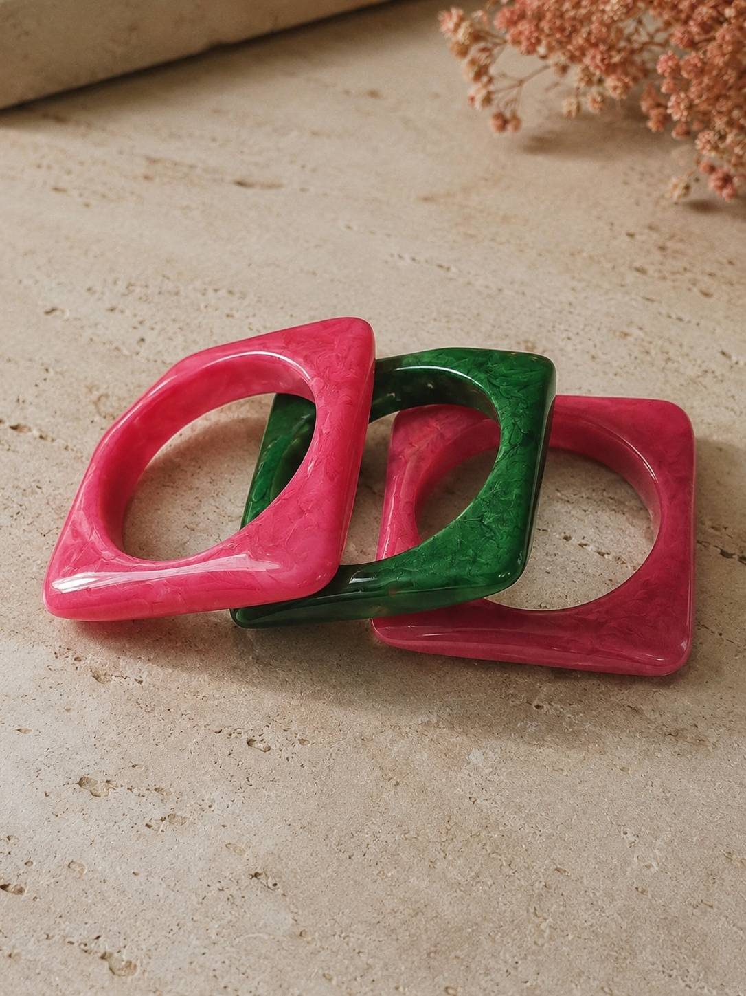 Three square marble-finish resin bangles in hot pink and emerald green arranged overlapping on a stone surface with dried flowers